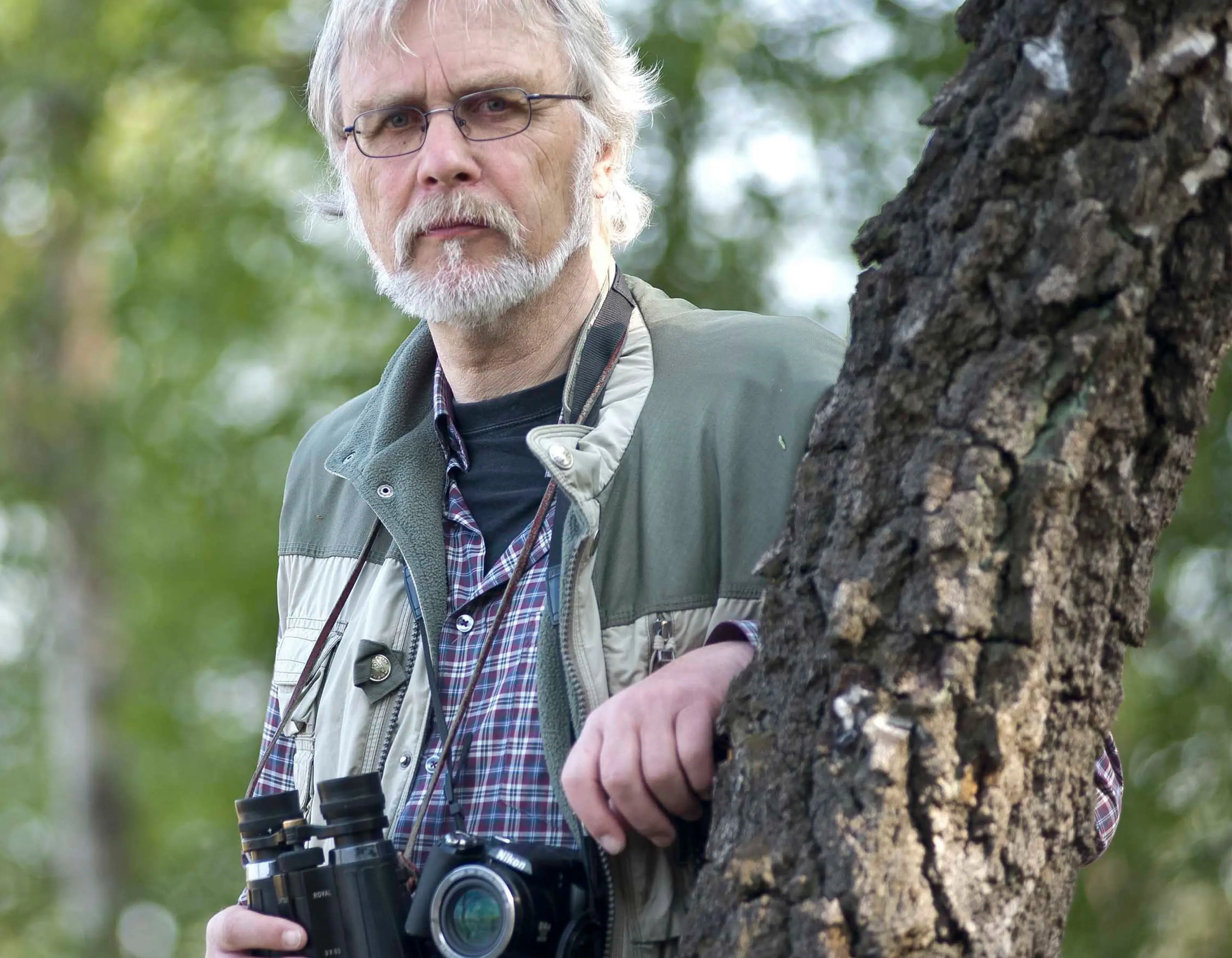 Ein älterer Mann mit grauem Haar, Brille und Bart steht im Freien neben einem Baumstamm. Er trägt eine grüne Outdoorjacke über einem karierten Hemd und hat ein Fernglas sowie eine Kamera um den Hals. Der Hintergrund zeigt grüne, unscharfe Bäume.