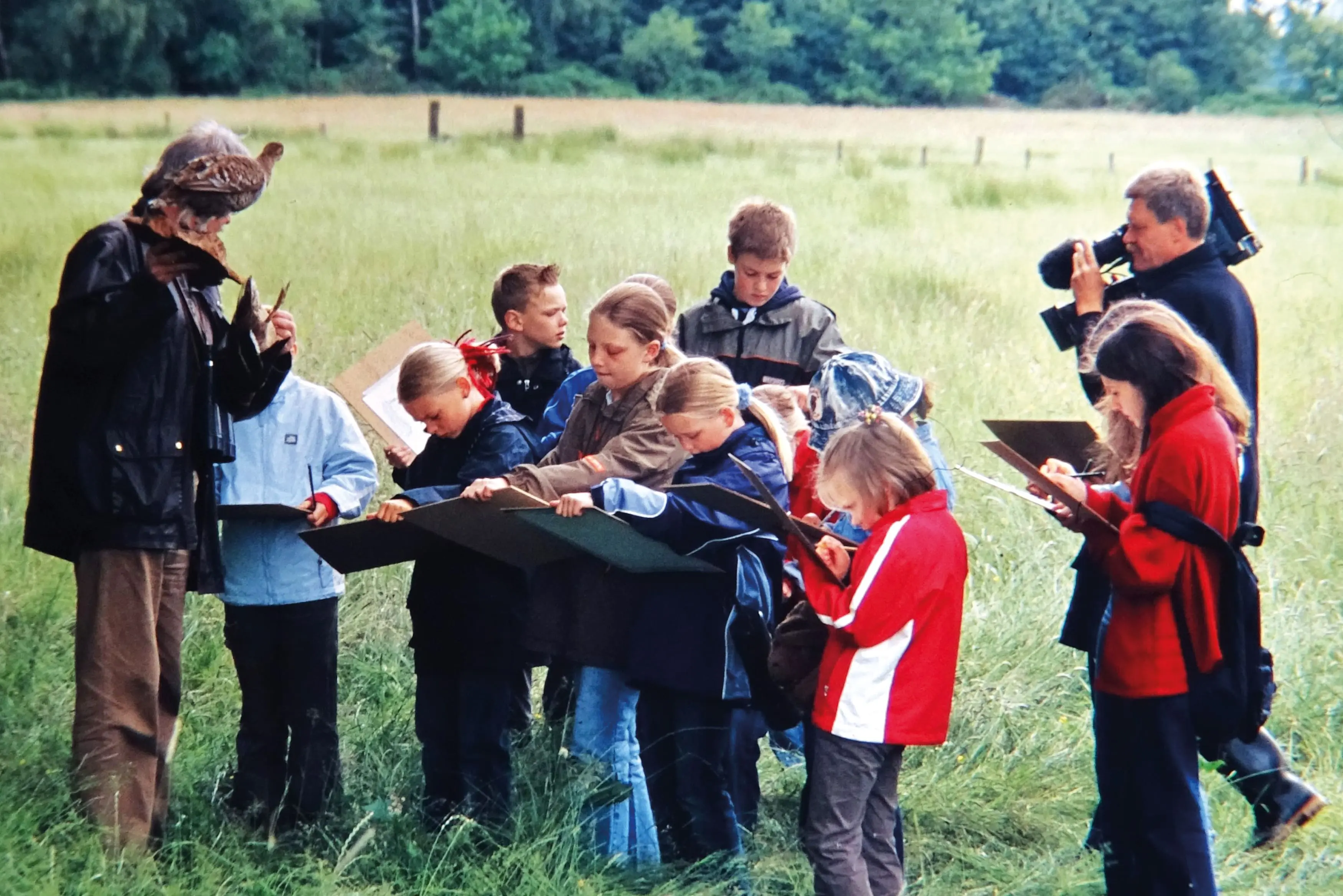 Mehrere Kinder, auf einer Wiese die einen Vogel Zeichnen. Hinter Ihnen steht ein Mann mit Kamera und vor Ihnen ein Mann mit einem Vogel auf dem Arm.