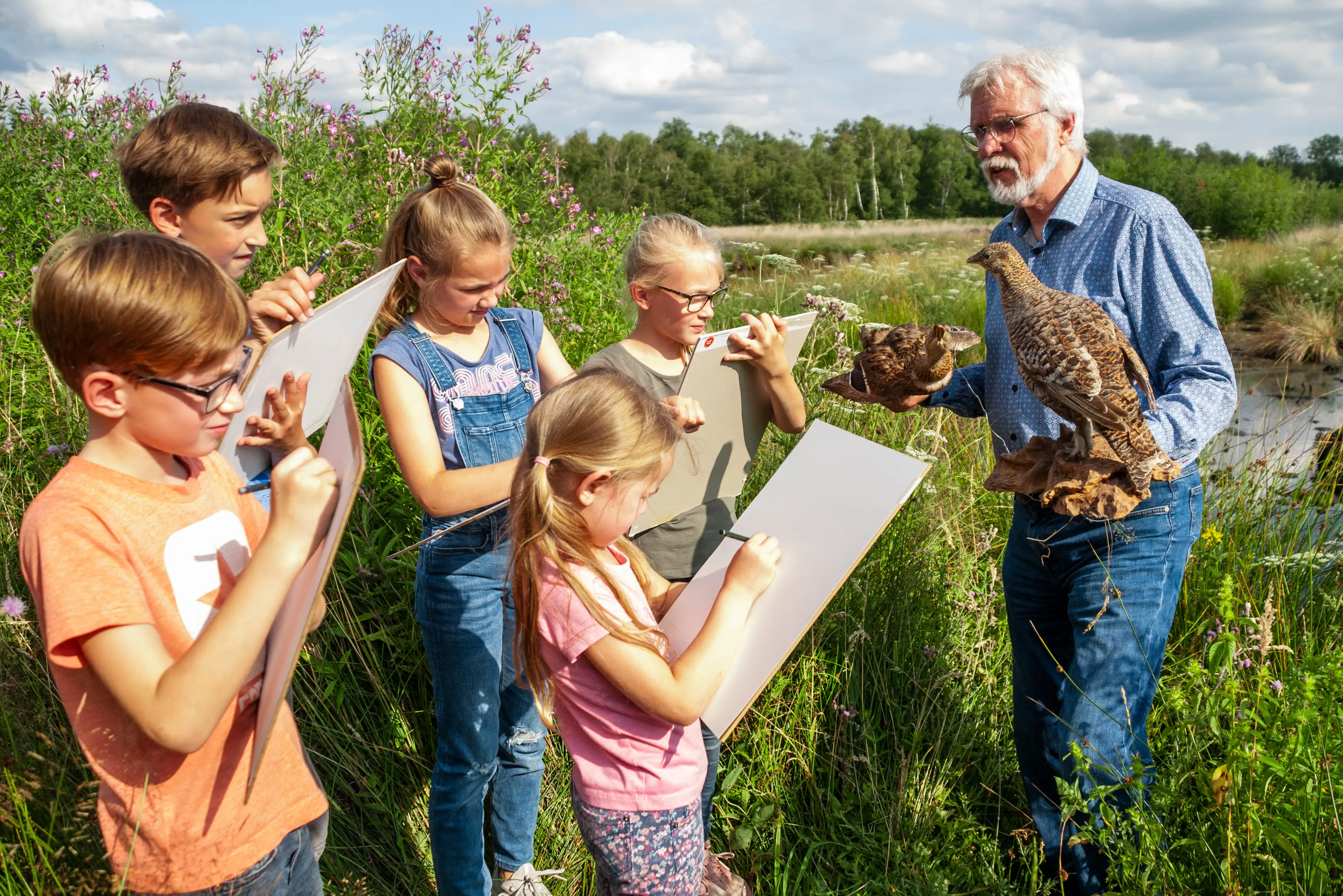 Fünf Kinder stehen auf einer Wiese, vor Ihnen steht ein Mann mit zwei Vögeln auf den Armen, die Kinder zeichnen diese Vögel.