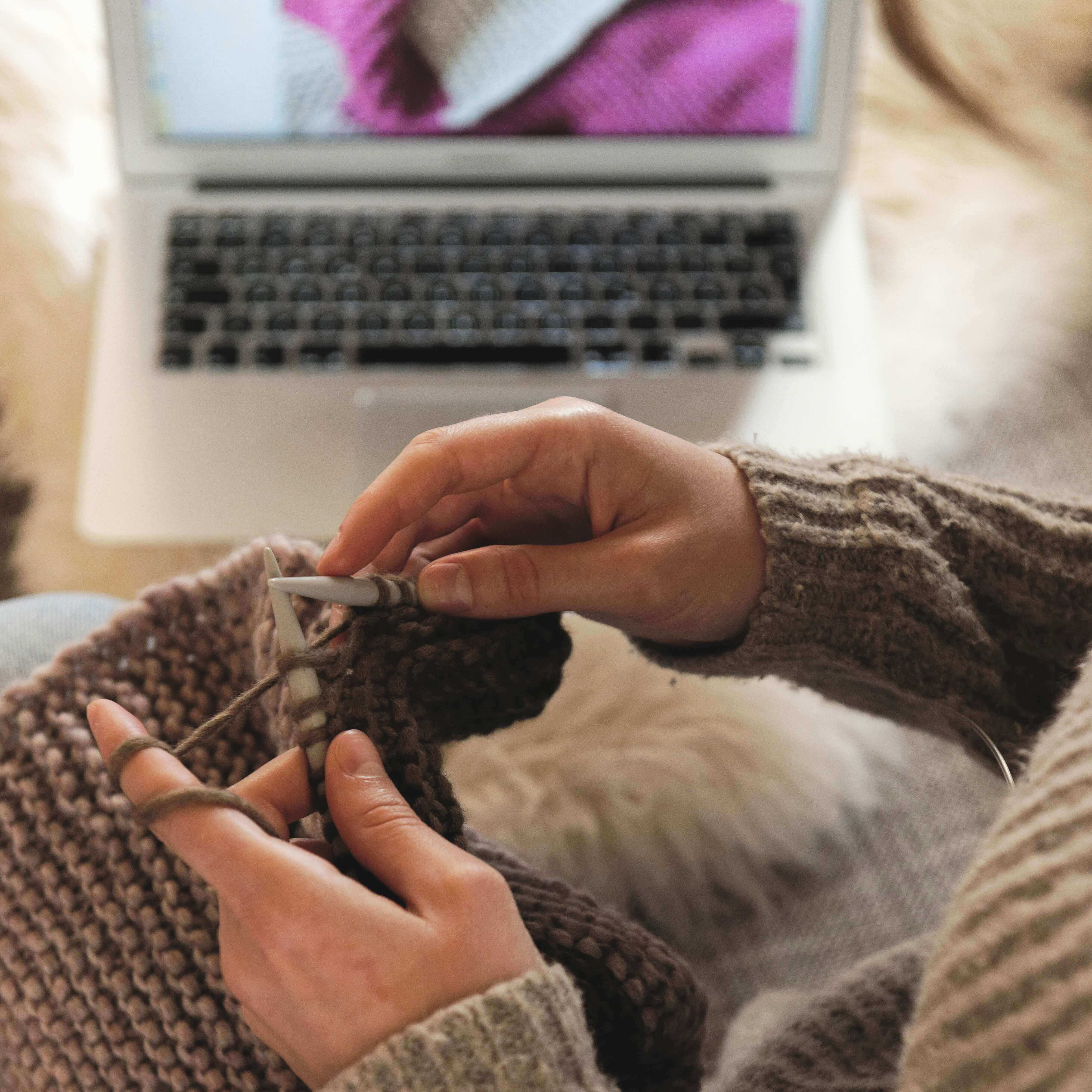 Person in grauem Pullover strickt mit brauner Wolle. Im Hintergrund ist ein Laptop mit einem Bild eines gestrickten Stücks auf dem Bildschirm sichtbar. Hände halten Stricknadeln und arbeiten an einem Projekt.