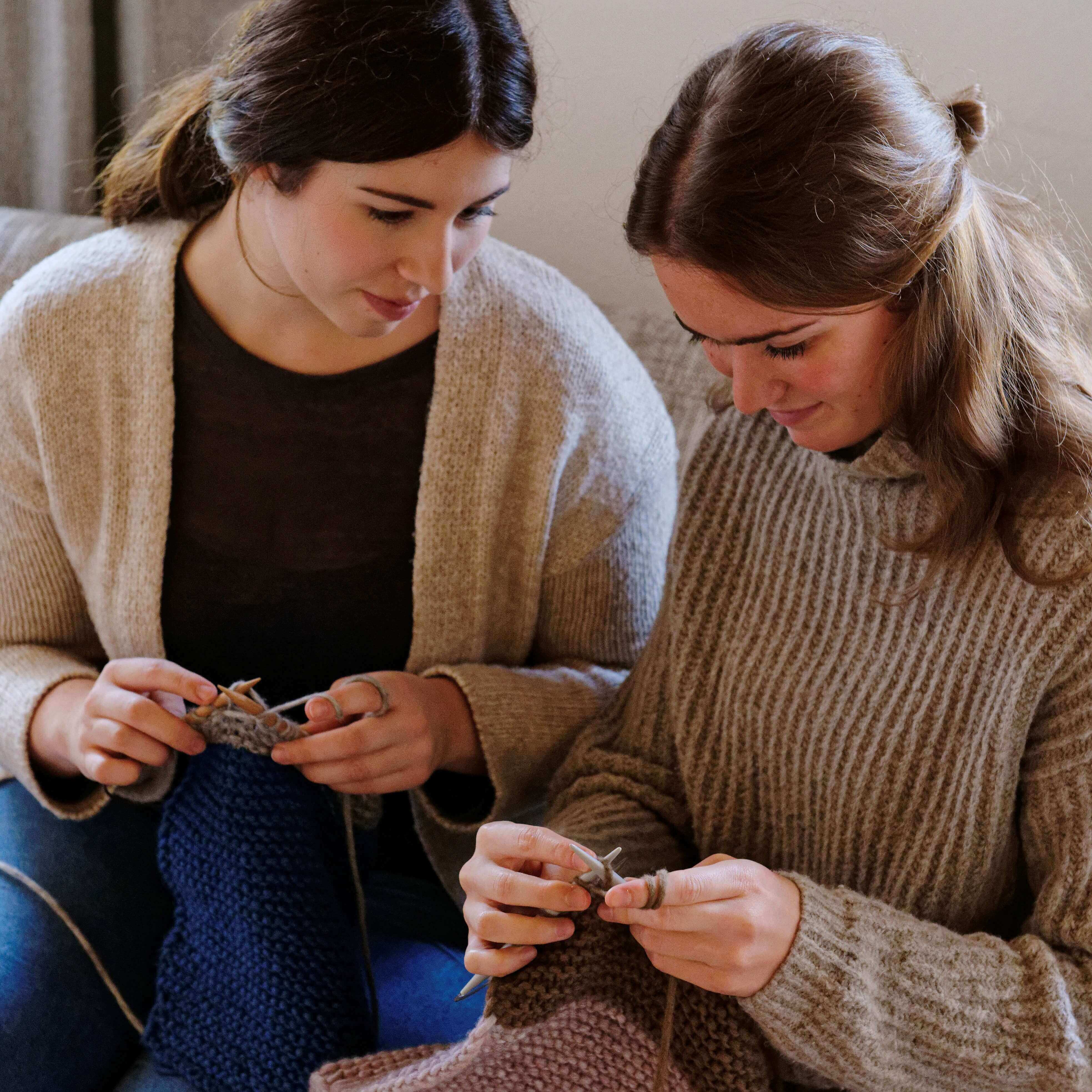 Zwei Frauen sitzen nebeneinander auf einem Sofa und stricken. Beide tragen Pullover. Die linke Frau hält ein blaues Strickstück, während die rechte Frau ein braunes Strickstück hält. Beide konzentrieren sich auf ihre Strickarbeit. 