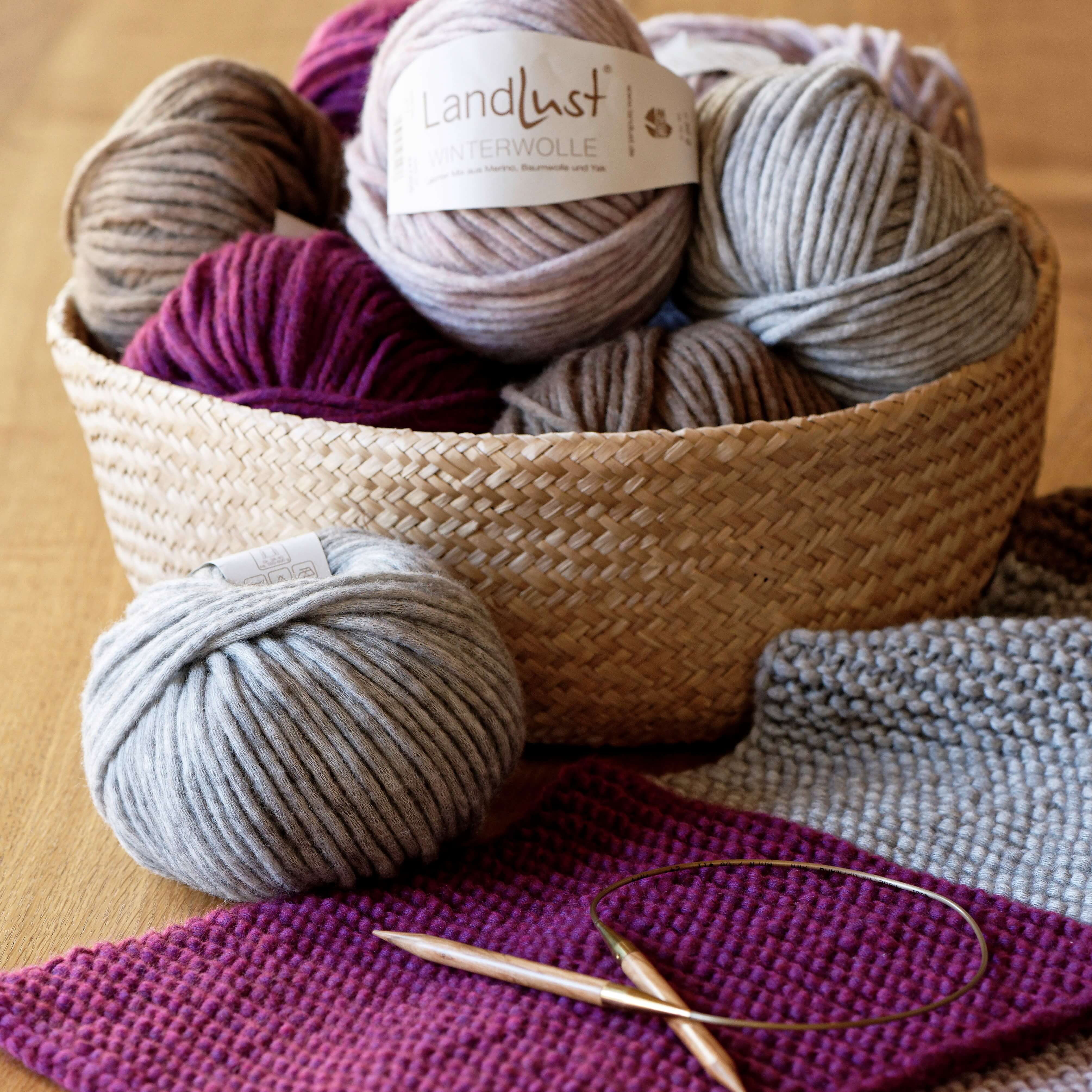 Ein geflochtener Korb mit bunten Wollknäueln, darunter grau, lila und beige, steht auf einem Holztisch. Vor dem Korb liegt ein graues und ein lila Strickstück mit zwei hölzernen Stricknadeln. Ein Wollknäuel trägt das Etikett „Landlust Winterwolle“.