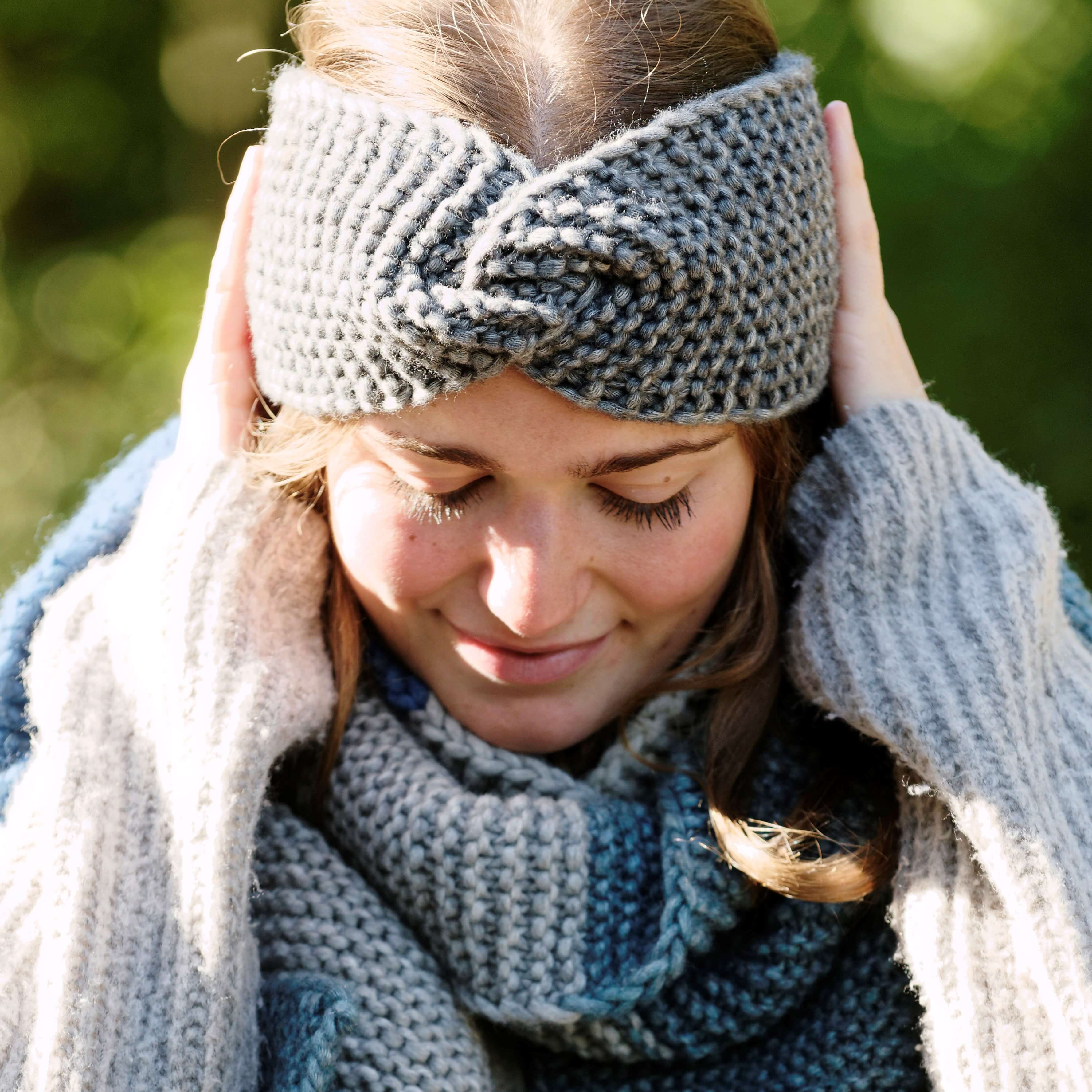 Frau trägt grauen, gestrickten Stirnband und Schal, die Hände an den Seiten des Kopfes haltend. Sie blickt nach unten mit geschlossenen Augen. Die Kleidung ist grob gestrickt. Der Hintergrund ist grün verschwommen, deutet auf eine Außenszene hin.