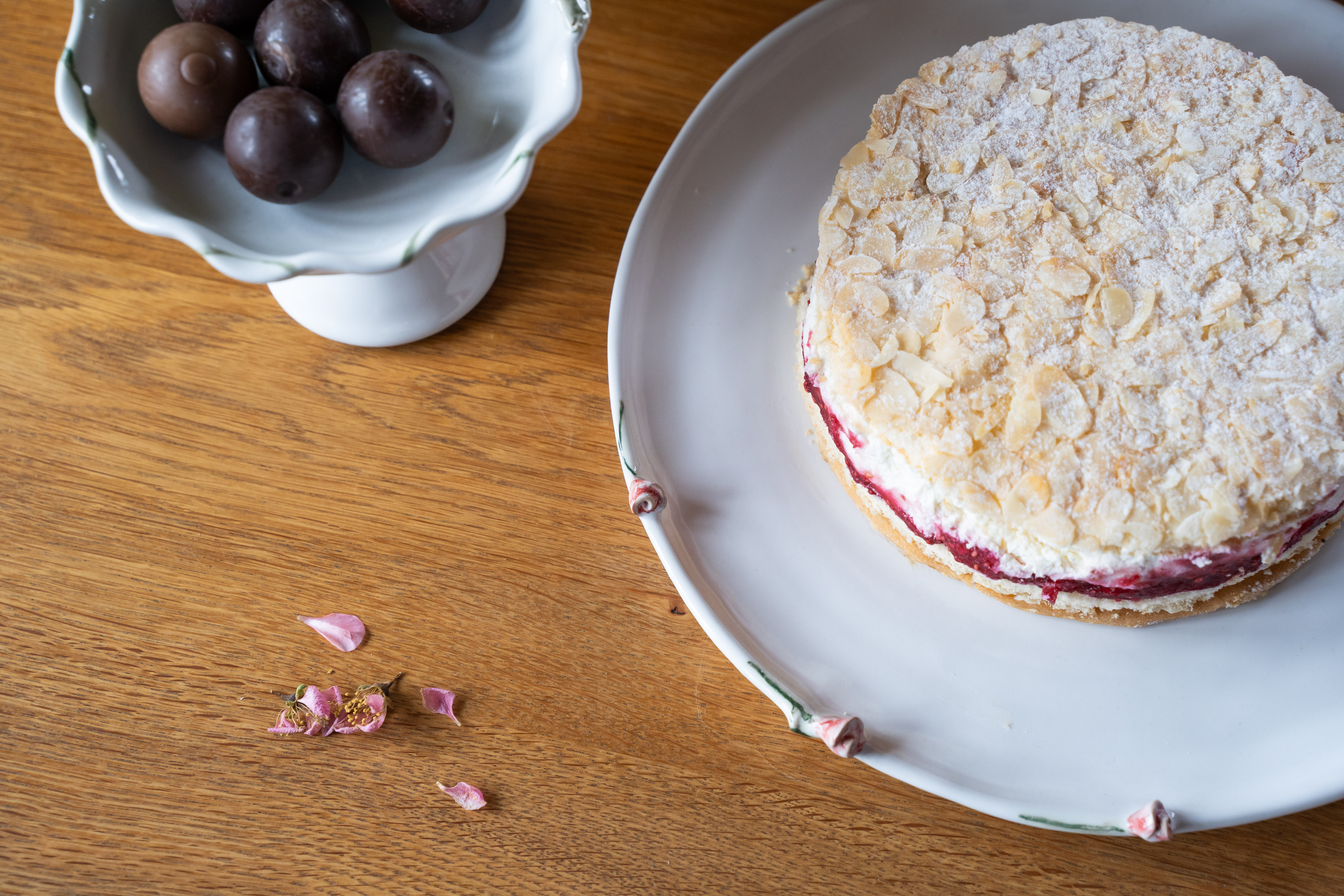 Ein Teller mit Mandelbaiser-Torte und rosafarbener Füllung, daneben Rosinenkugeln in einer Schale; auf einem Holztisch liegen getrocknete Blütenblätter.