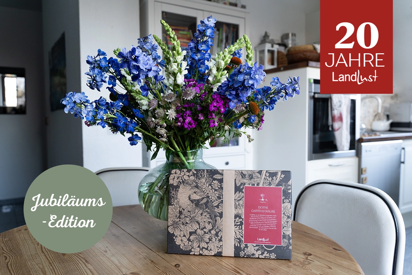 Großer Blumenstrauß in Vase auf Holztisch, daneben Buch mit floralem Mustercover. Im Hintergrund Küche. Oben rechts rotes Feld mit weißem Text 20 Jahre Landlust. Grüner Kreis links mit weißem Text Jubiläums-Edition. Bild verlinkt zum Produkt.