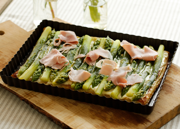 Rechteckige Quiche mit grünem Spargel und Schinkenstreifen, auf einem Holzbrett serviert. Im Hintergrund zwei Vasen mit grünen Blättern. Bild verlinkt zum Rezept