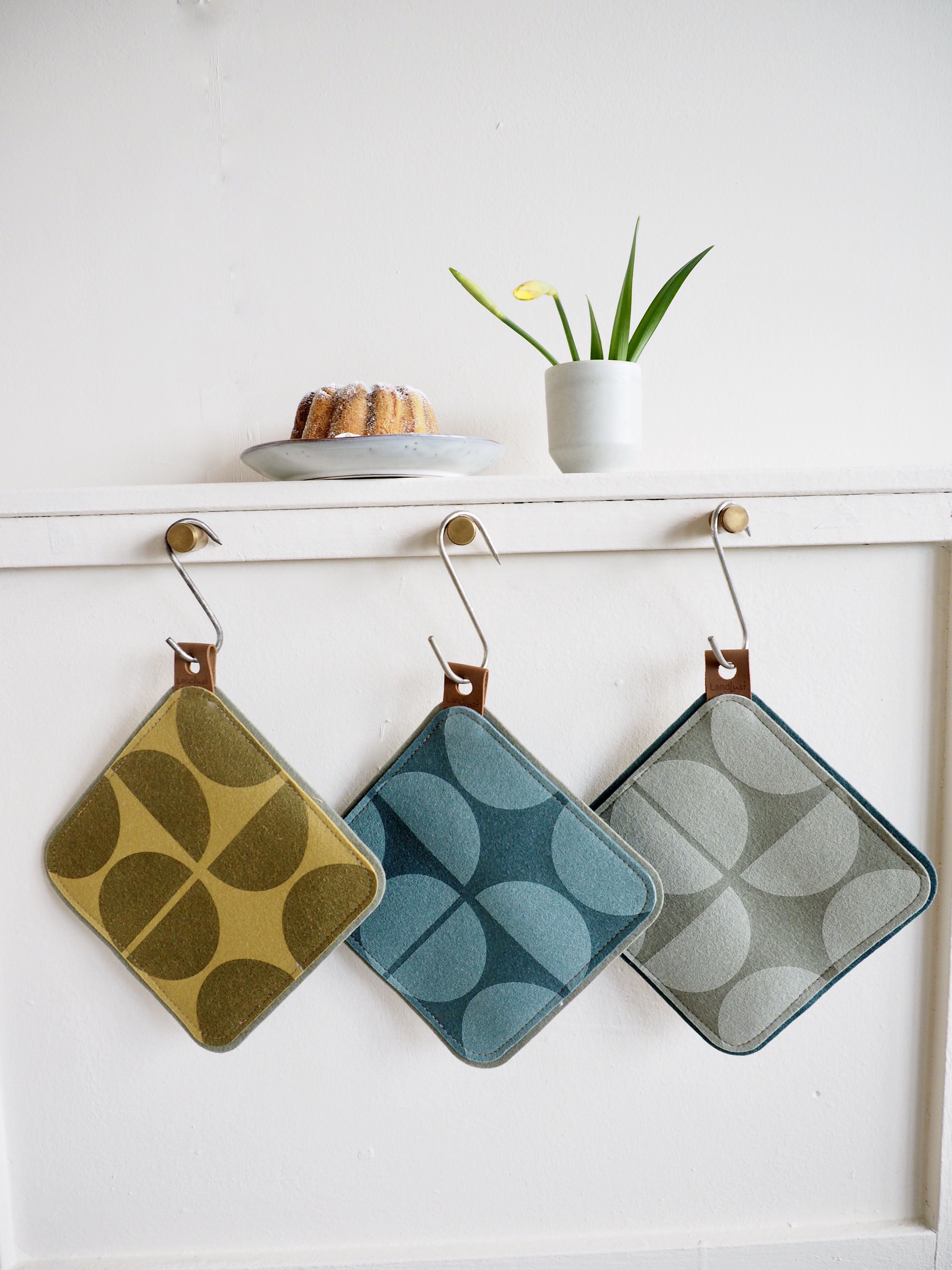 Drei quadratische Topflappen in verschiedenen Farben hängen nebeneinander an Haken an einer weißen Wand. Oberhalb steht ein Teller mit einem kleinen Kuchen neben einer weißen Tasse mit einer grünen Pflanze. Bild verlinkt zum Produkt