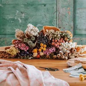 Ein Holztisch mit mehreren kleinen, in braunes Papier gewickelten Sträußen aus getrockneten Blumen in verschiedenen Farben, darunter Gelb, Rosa, Weiß und Lila. Im Hintergrund eine grüne, abgenutzte Holzwand. Auf dem Tisch liegt eine Stoffdecke.