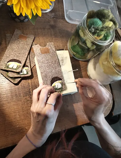 Nahaufnahme von Händen, die mit einer Filznadel farbige Wollfasern in ein Etui aus Wollfilz einarbeiten.