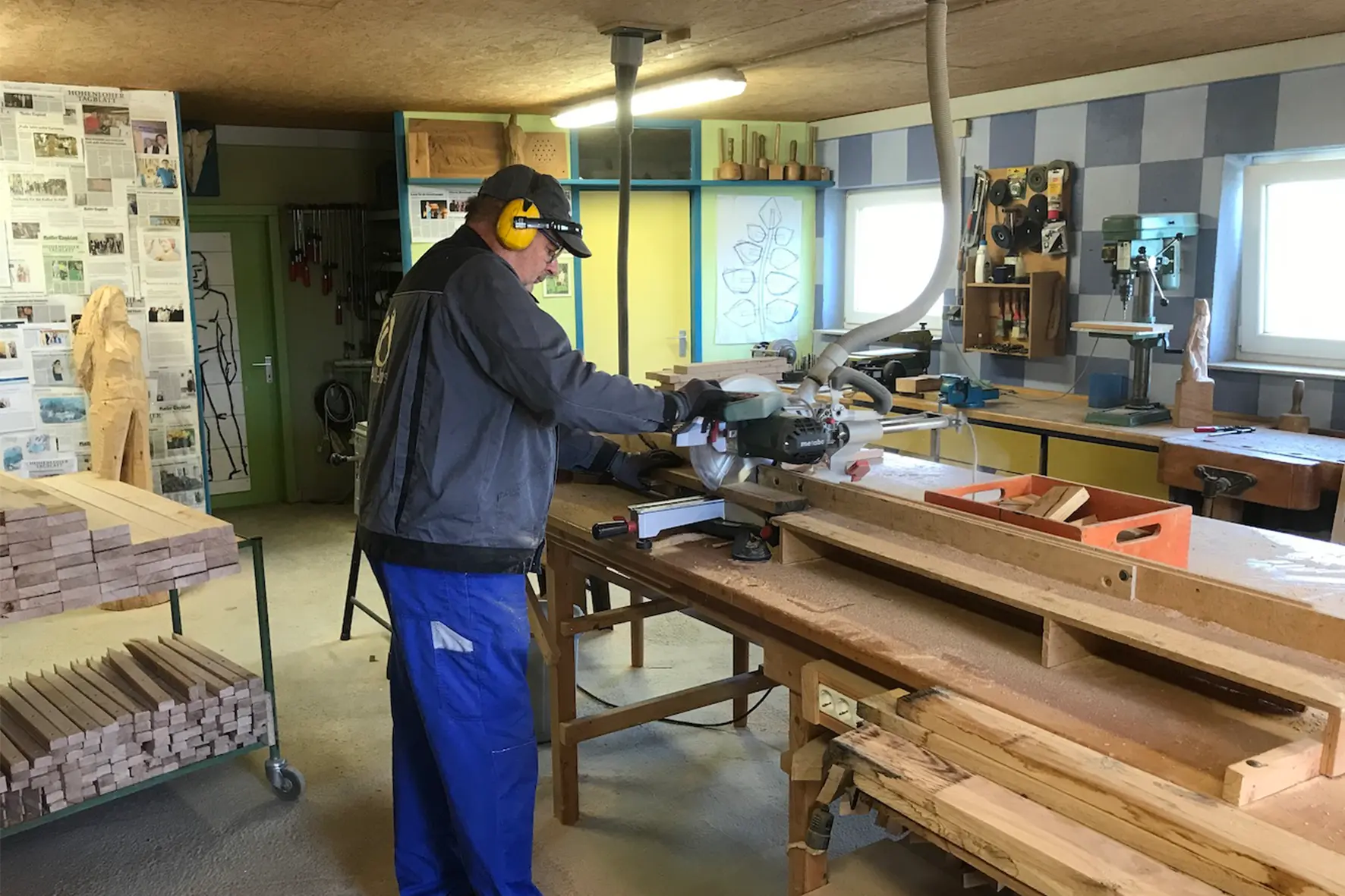 Ein Mann in Schutzkleidung arbeitet in einer hellen Holzwerkstatt an einer Tischkreissäge, umgeben von Holzstücken und Werkzeugen.