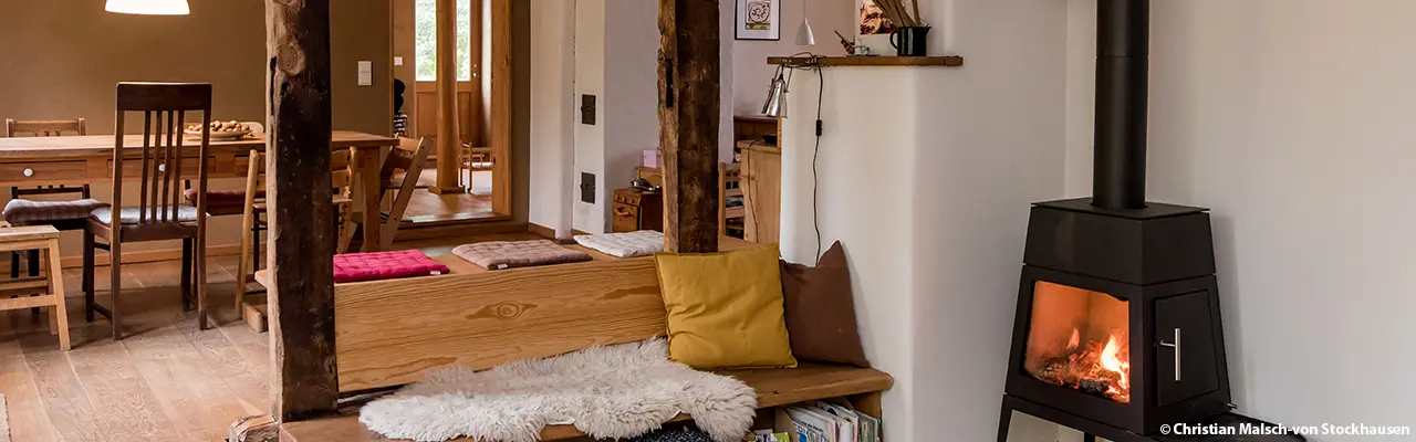 Gemütliches Wohnzimmer mit Holzmöbeln, Esstisch und Stühlen im Hintergrund. Sitzbank mit bunten Kissen und Fell, neben einem schwarzen Kaminofen mit brennendem Feuer, sichtbare Holzbalken trennen die Bereiche.