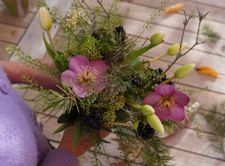 Eine Person in lila Pullover hält einen Blumenstrauß aus rosa Blüten, grünen Blättern, kleinen gelben Knospen und dunklen Beeren. Der Strauß liegt auf einem hölzernen Tisch mit verstreuten Blütenblättern. Bild verlinkt zum Workshop.