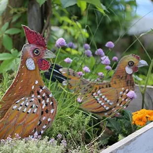 Zwei bemalte Hühnerfiguren aus Holz stehen inmitten von blühenden lilafarbenen und orangefarbenen Blumen in einem Pflanzenbeet. Im Hintergrund sind grüne Blätter und ein unscharfer Himmel erkennbar. Bild verlinkt zum Workshop.