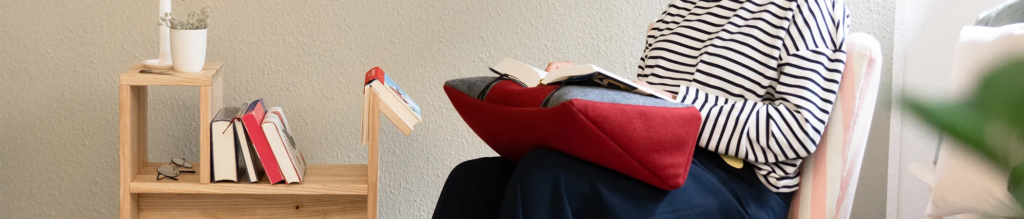 Person sitzt auf einem Stuhl und liest ein Buch auf einem roten Kissen mit Buchstütze auf den Knien. Neben ihr steht ein Holztisch mit mehreren Büchern, einer umgedrehten Brille und einer kleinen Topfpflanze.