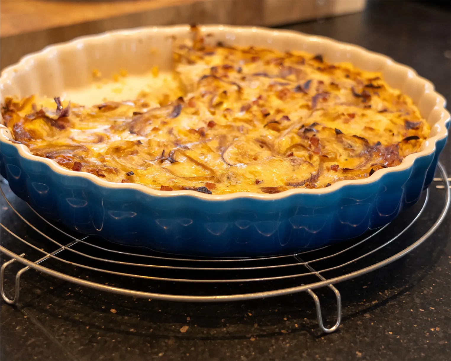 Eine blaue Keramik-Backform auf einem Metallgitter zeigt einen frisch gebackenen Zwiebelkuchen mit goldbraun gebräunten Zwiebeln und Käse überzogen. Die Backform steht auf einer dunklen Küchenarbeitsplatte. Bild verlinkt zum Workshop