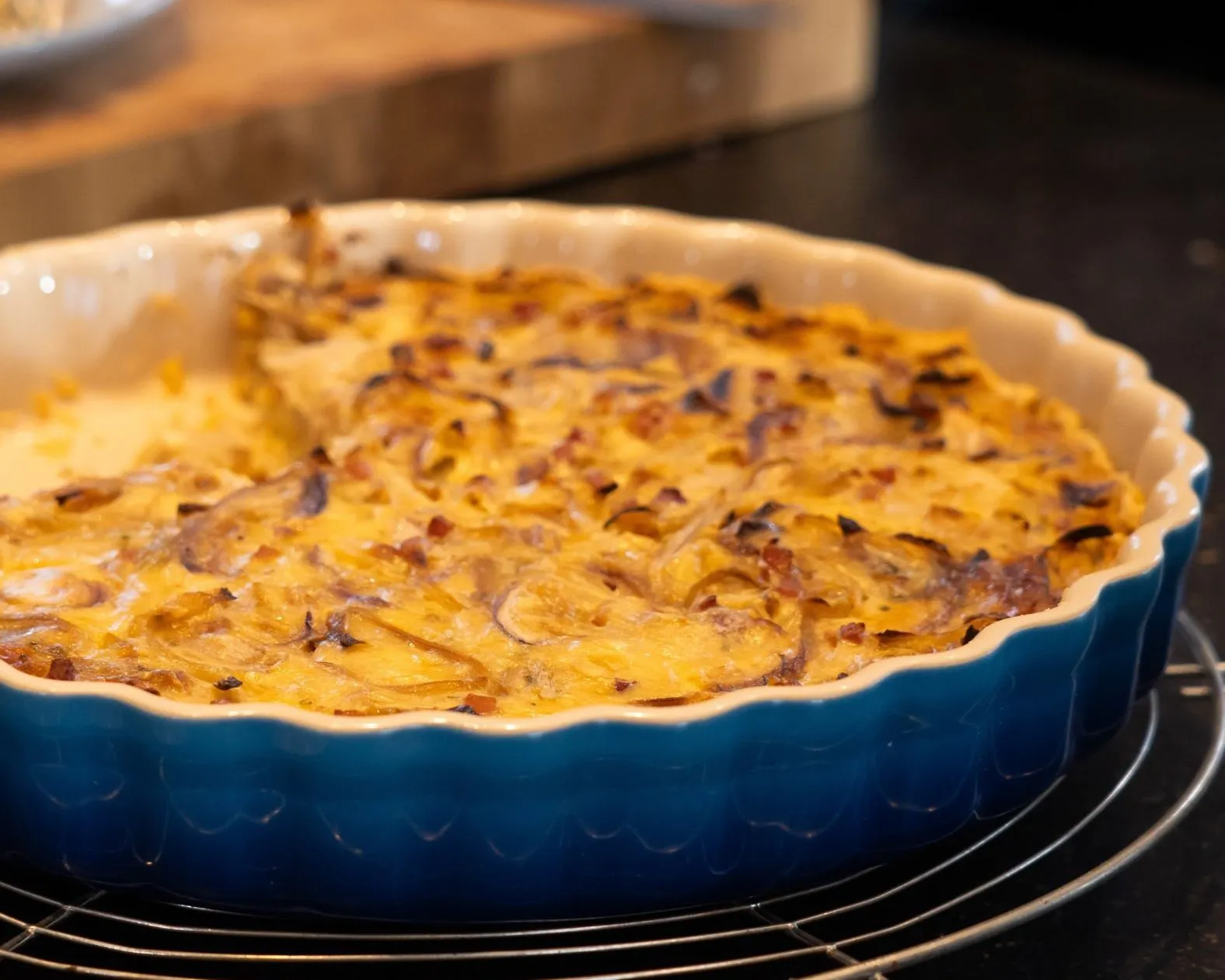 Eine runde, blaue Auflaufform mit einer angeschnittenen Zwiebelkuchen in goldbrauner Kruste. Der Kuchen enthält sichtbare Zwiebel- und Speckstücke. Die Form steht auf einem runden Metallgitter. Im Hintergrund ist ein Holzbrett. Bild verlinkt zum Workshop.