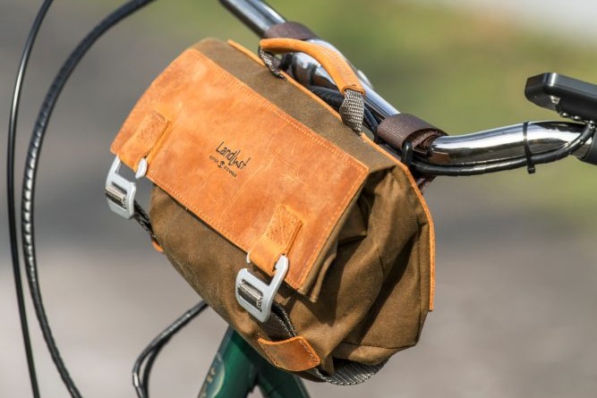 Eine braune Fahrradtasche aus Leder und Stoff ist am Lenker eines Fahrrads befestigt. Die Tasche hat silberne Schnallenverschlüsse und ein kleines Logo in der Mitte des Deckels, Bild verlinkt zum Produkt.