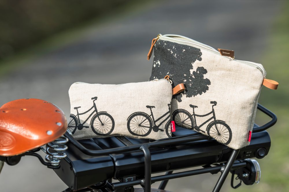 Zwei cremefarbene Taschen mit schwarzen Fahrradmotiven liegen auf einem Fahrradgepäckträger. Die eine Tasche zeigt ein vollständiges Fahrrad, die andere nur den schwarzen Umriss. Beide Taschen haben braune Reißverschlüsse. Bild verlinkt zum Produkt.
