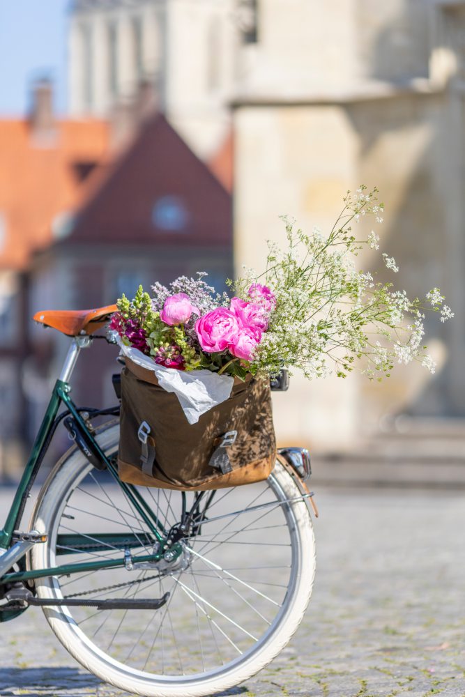 Ein Fahrrad mit einem braunen Korb am hinteren Gepäckträger. Der Korb ist gefüllt mit rosa und weißen Blumen. Im Hintergrund sind unscharf Gebäude aus hellem Stein und rote Ziegeldächer zu sehen.