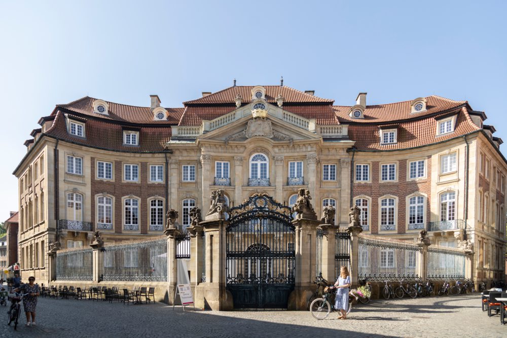 Ein großes, historisches Gebäude mit einem geschwungenen, zweistöckigen Barockstil, roten Dächern und zahlreichen Fenstern. Der Eingangsbereich ist mit einem verzierten schmiedeeisernen Tor und Statuen geschmückt. Eine Person mit Fahrrad steht rechts.