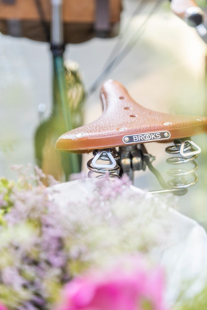 Ein brauner Fahrradsattel befindet sich im Hintergrund, teilweise verdeckt durch blühende Blumen im Vordergrund. Der Rest des Bildes ist verschwommen.