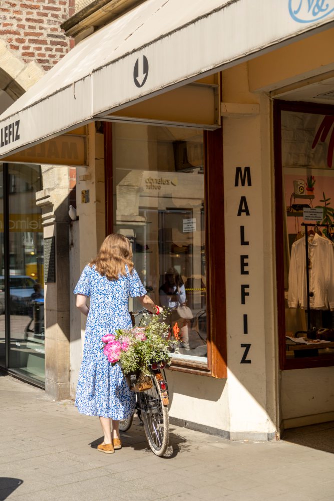 Eine Person in einem blauen Kleid schiebt ein Fahrrad mit einem Korb voller Blumen vor einem Geschäft mit der Aufschrift „MALEFIZ“ am Schaufenster. Die Ladenfassade ist beige, mit großen Schaufenstern, die verschiedene Dekorationsartikel zeigen.