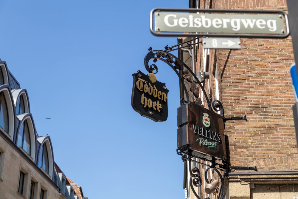 Straßenschild „Gelsbergweg“ an der Ecke eines Backsteingebäudes. Darunter zwei hängende Schilder: Eines mit der Aufschrift „Gösser Bier“ und das andere mit einem Wappen und der Aufschrift „Rittmayer Private Brauerei“. 