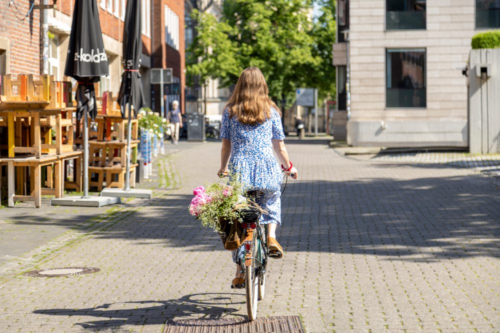 Eine Frau in einem blauen Kleid fährt bei sonnigem Wetter auf einem Fahrrad durch eine gepflasterte Straße. Der Fahrradkorb ist mit bunten Blumen gefüllt. Links befinden sich Tische und Stühle vor einem Gebäude mit schwarzen Sonnenschirmen. 