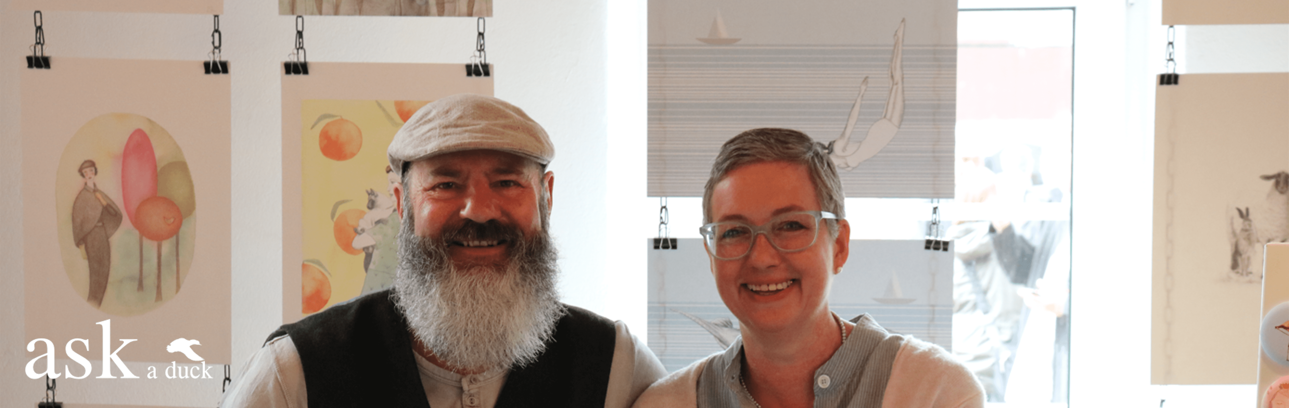 Header zur Landingpage As a Duck: Fotografie eines Paares, bestehend aus einer Frau mit kurzen Haaren und einem Mann mit Bart, beide lächelnd, vor einem neutralen Hintergrund.