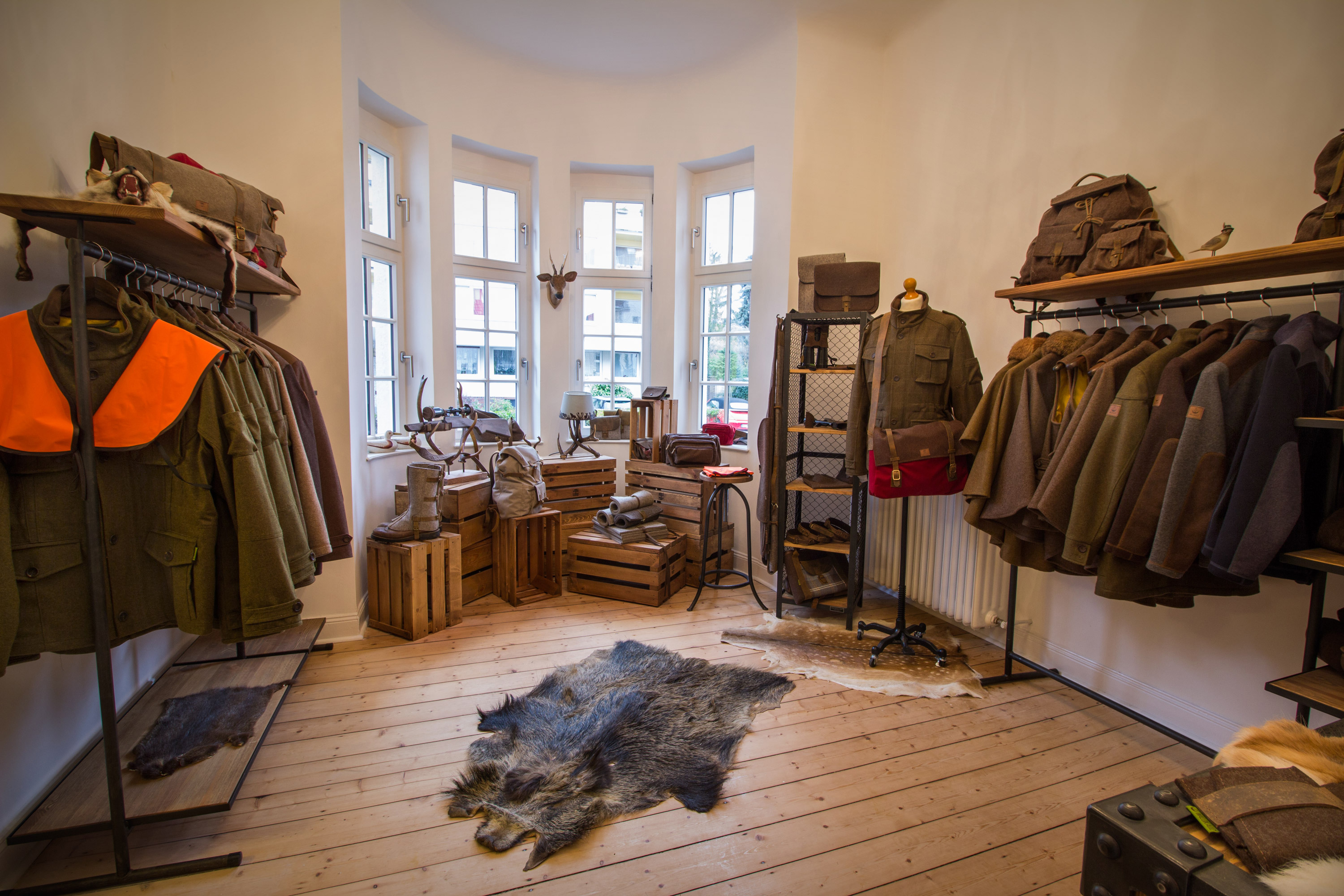 Ein kleiner Laden mit Holzboden und weißen Wänden zeigt Outdoor-Bekleidung auf Kleiderständern. Im Hintergrund stehen Holzkisten und ein Tisch mit Accessoires vor großen Sprossenfenstern. Tierfell liegt auf dem Boden.