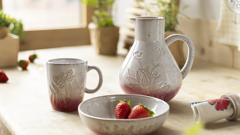 Keramikgeschirr-Set mit Kanne, Tasse und Schale mit Blumenmotiv, auf Holztisch platziert. Drei frische Erdbeeren in der Schale, umgeben von Pflanzen im Hintergrund, leichte Unschärfe.