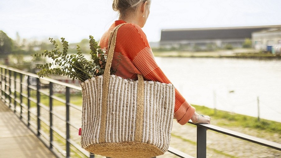 Frau in orangefarbenem Oberteil steht mit gestreifter Strohtasche voller Blumen am Geländer eines Flusses, Blick aufs Wasser gerichtet. Bild verlinkt das Produkt.