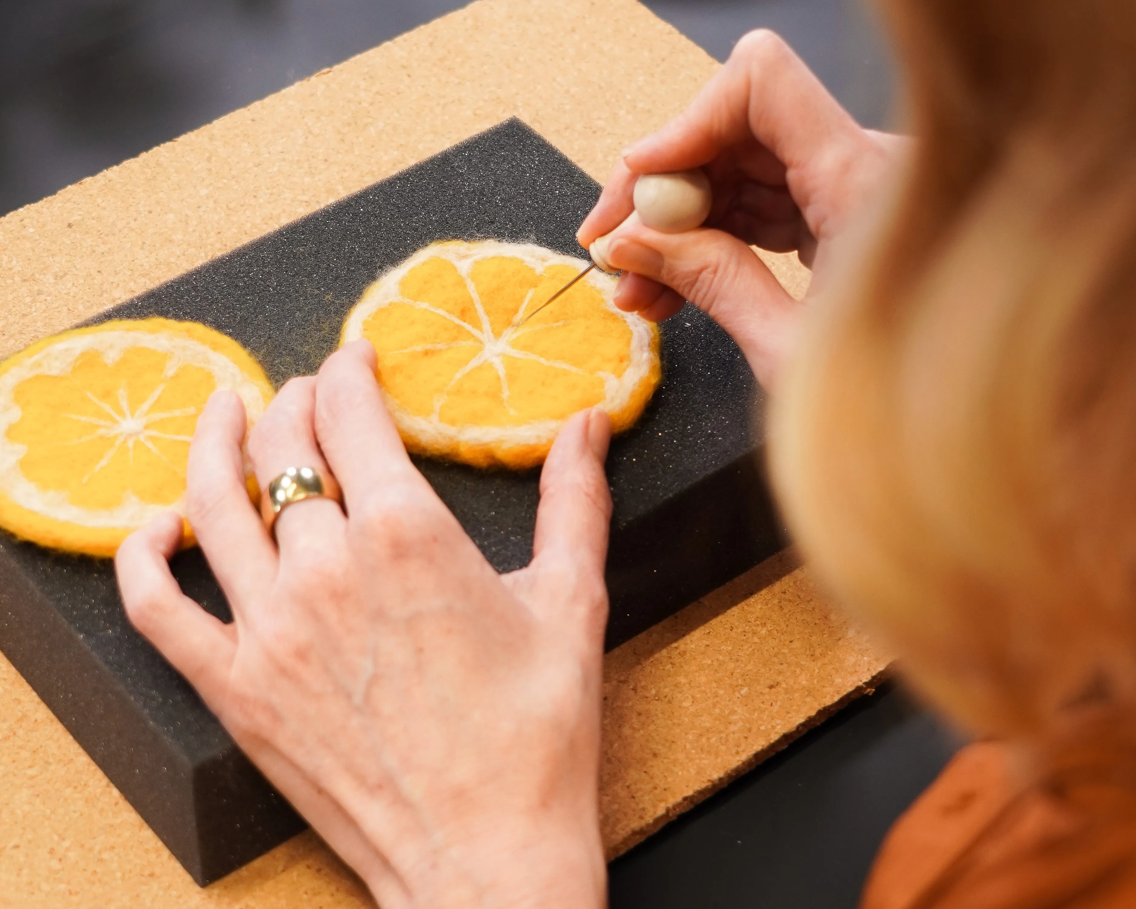 Person schnitzt mit einem Werkzeug ein Muster in eine Orangenscheibe, die auf einem dunklen Kissen aufliegt. Eine zweite Orangenscheibe liegt daneben. Bild verlinkt zum Workshop