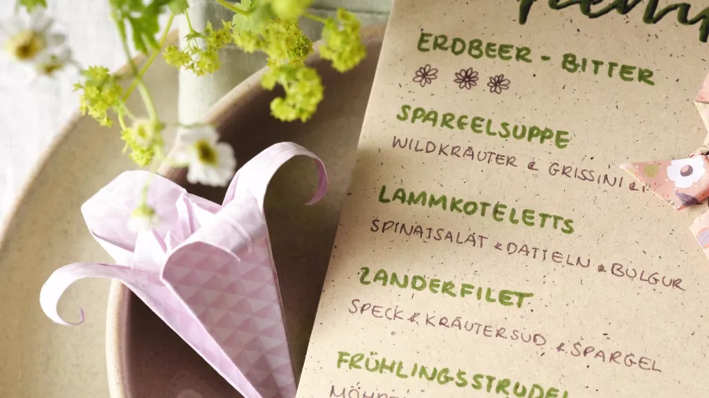 Handgeschriebenes Menü auf braunem Papier neben rosa gefaltetem Stoffservietten-Halter in Blumenform. Menü enthält: Erdbeer-Bitter, Spargelsuppe mit Wildkräutern und Grissini, Lammkoteletts mit Spinatsalat, Datteln und Bulgur Bild verlinkt zur Anleitung. 