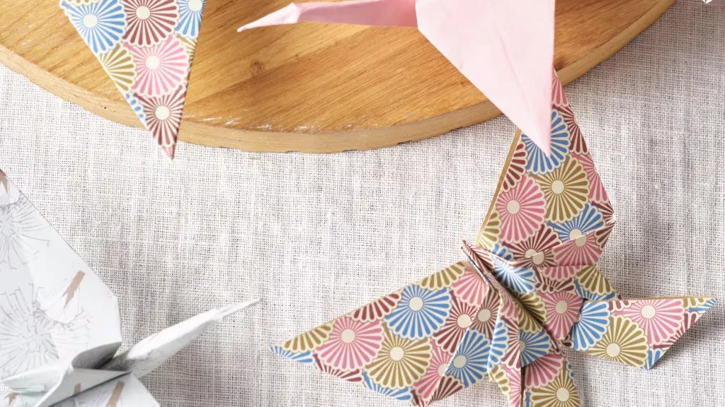 Farbige Origami-Figuren, einschließlich eines rosa Kranichs und eines gemusterten Schmetterlings mit Blumenmuster in Blau und Rosa, liegen auf einem Leinentuch. Im Hintergrund ist ein rundes Holztablett zu sehen. Bild verlinkt zur Anleitung.