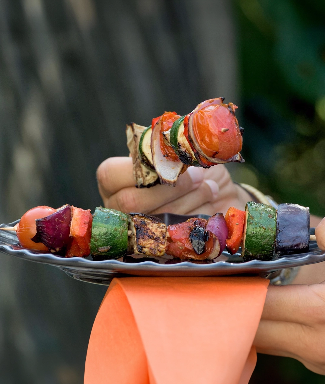 Ein Teller mit gegrillten Gemüsespießen wird von einer Hand gehalten. Die Spieße enthalten Zucchini, Paprika, Zwiebeln und Tomaten. Eine orangefarbene Serviette wird unter dem Teller gehalten.
