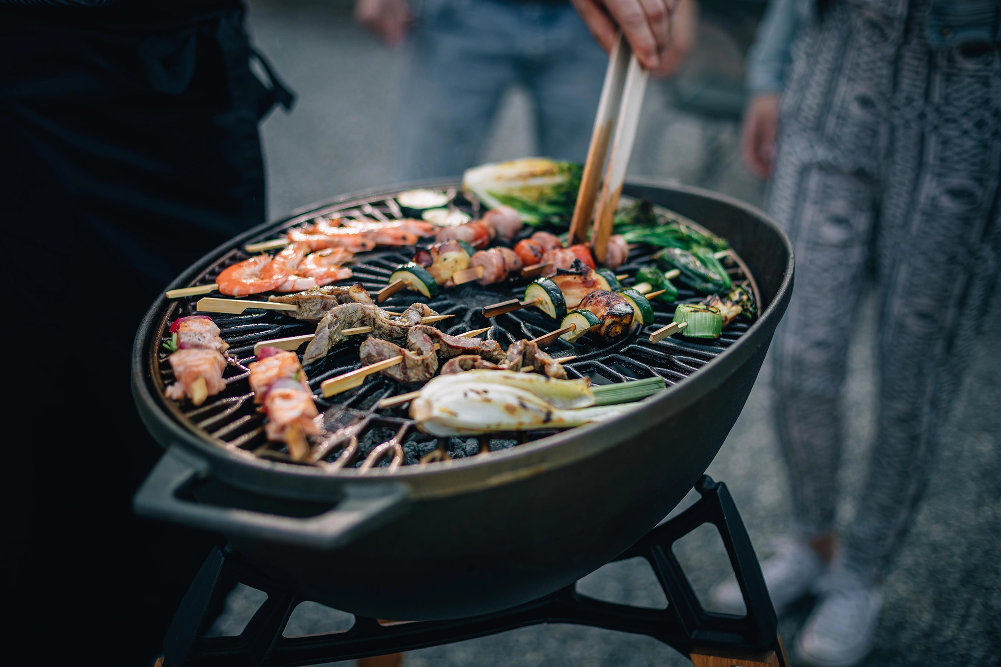 Ein runder Grill mit offenem Deckel, auf dem Gemüse, Fleischspieße und Maiskolben brutzeln. Eine Hand hält eine Grillzange und wendet das Essen, Bild verlinkt zum Produkt.