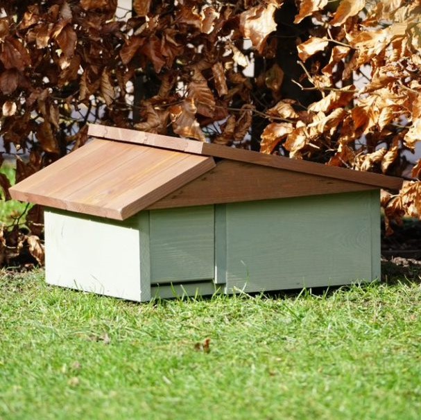 Holz-Igelhaus mit schrägem, braunem Dach und grünlichen Wänden, platziert auf grünem Gras vor einer Hecke mit trockenem Laub.