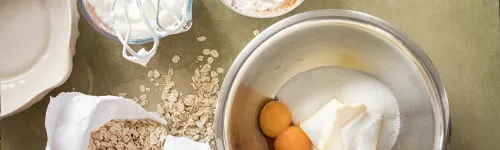 Schüssel mit Zucker, Butter und zwei Eigelb. Daneben eine angebrochene Papiertüte mit Haferflocken, einige Flocken auf einem Tisch ausgebreitet. Ein leerer weißer Teller liegt links oben. Der Hintergrund ist beige.