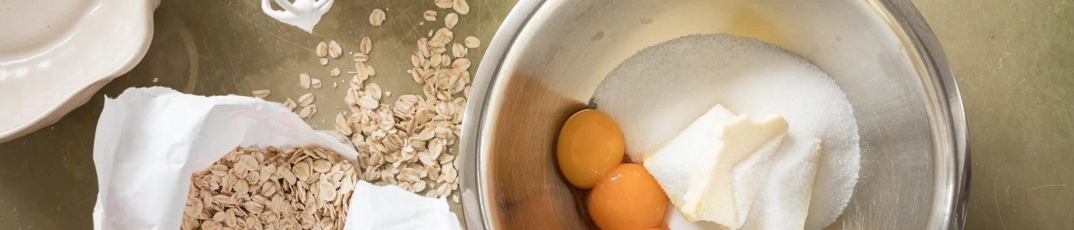 Schüssel mit Zucker, Butter und zwei Eigelb. Daneben eine angebrochene Papiertüte mit Haferflocken, einige Flocken auf einem Tisch ausgebreitet. Ein leerer weißer Teller liegt links oben. Der Hintergrund ist beige.