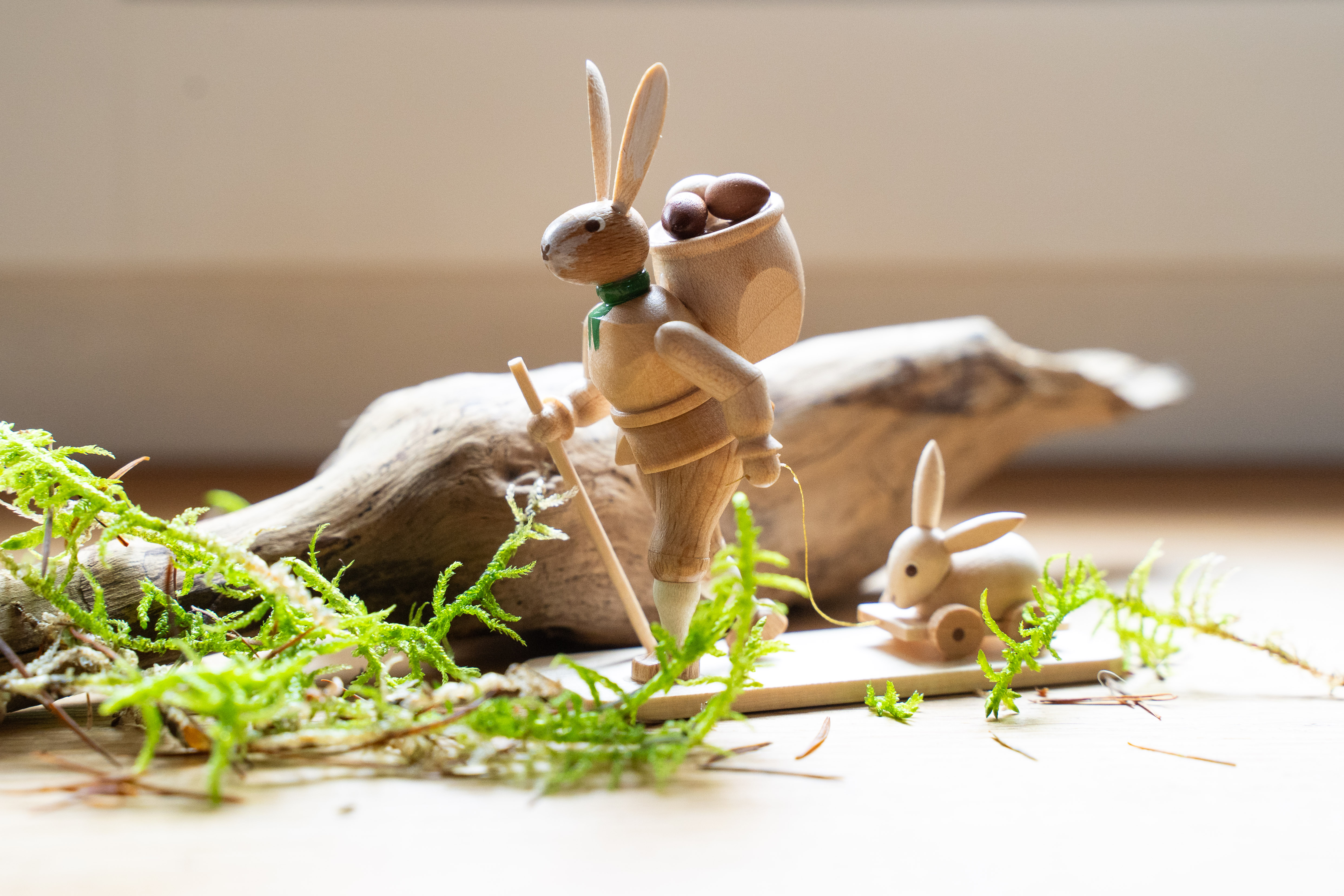 Holzfiguren eines Hasen mit Stock und einem kleineren Hasenkind auf Holzblock, umgeben von Moos und einem Holzstück, vor neutralem Hintergrund.
