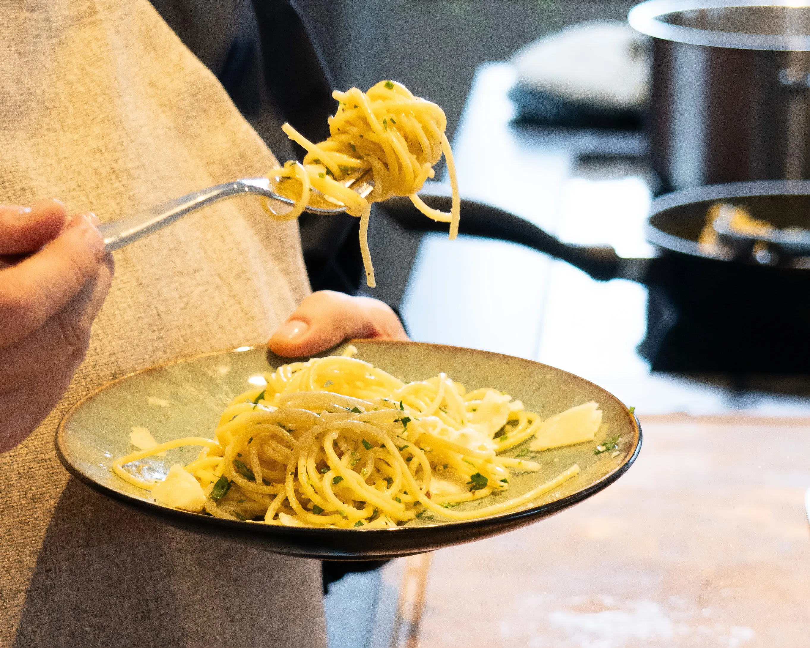 Person hält einen Teller mit Spaghetti in der linken Hand, aufgeteilt mit einer Gabel in der rechten Hand. Die Spaghetti sind mit Kräutern und Käsestückchen garniert. Im Hintergrund sind verschwommene Küchengeräte sichtbar. Bild verlinkt zum Workshop.