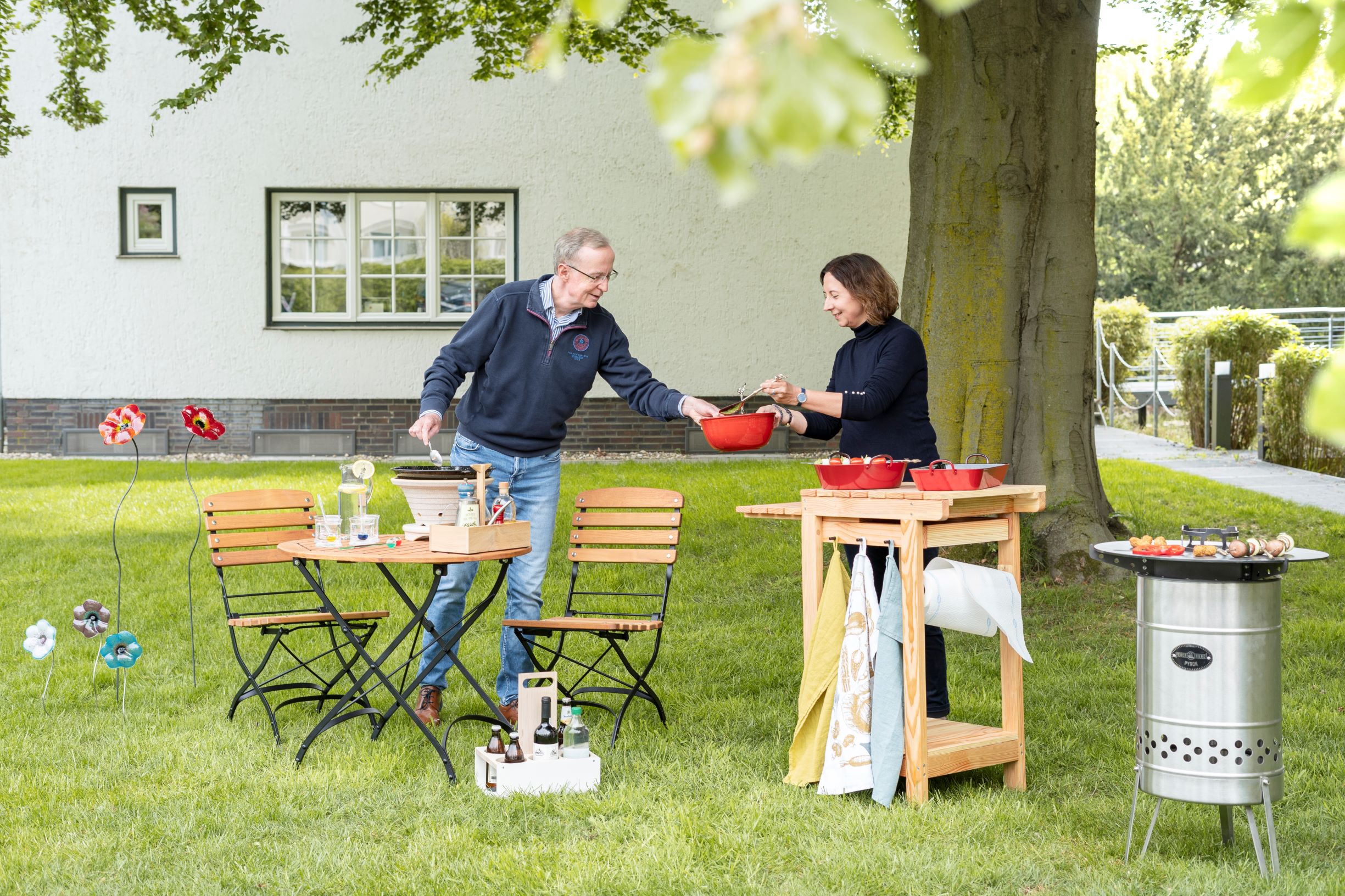 Ein älterer Mann und eine Frau stehen im Garten vor einem weißen Haus. Der Mann hält eine Flasche in der Hand, während die Frau eine rote Schüssel auf einen Holztisch stellt. Neben ihnen steht ein runder Tisch mit zwei Stühlen. Bild verlinkt zu Produkten