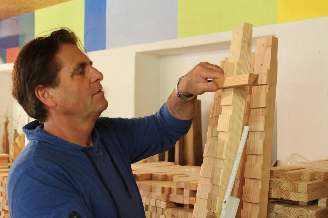Ein Mann in blauem Pullover arbeitet konzentriert an einem Holzturm mit einem Winkelmaß in der Hand, umgeben von Holzblöcken. Der Hintergrund zeigt eine bunte Wand.