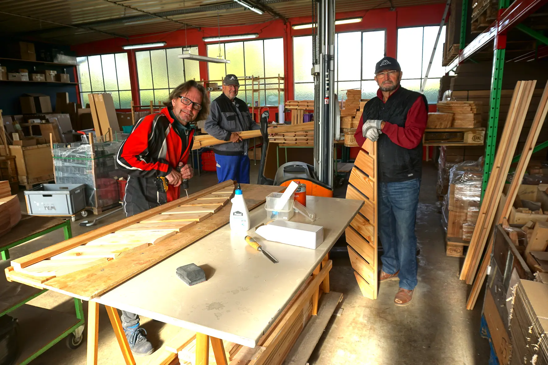 Drei Männer arbeiten in einer Werkstatt. Zwei von ihnen sägen Holz an einer Tischkreissäge. Der dritte Mann hält ein dreieckiges Holzstück. Im Hintergrund sind Regale mit Werkzeugen und Holzmaterialien sichtbar.
