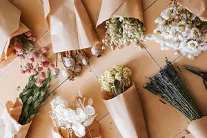 Mehrere Blumensträuße in braunem Papier eingewickelt liegen in einem Rastermuster auf einer hellen, fliesenähnlichen Oberfläche. Zu den sichtbaren Blumen gehören rosafarbene und weiße Blüten und gelbe Knospen.  Bild verlinkt zum Produkt