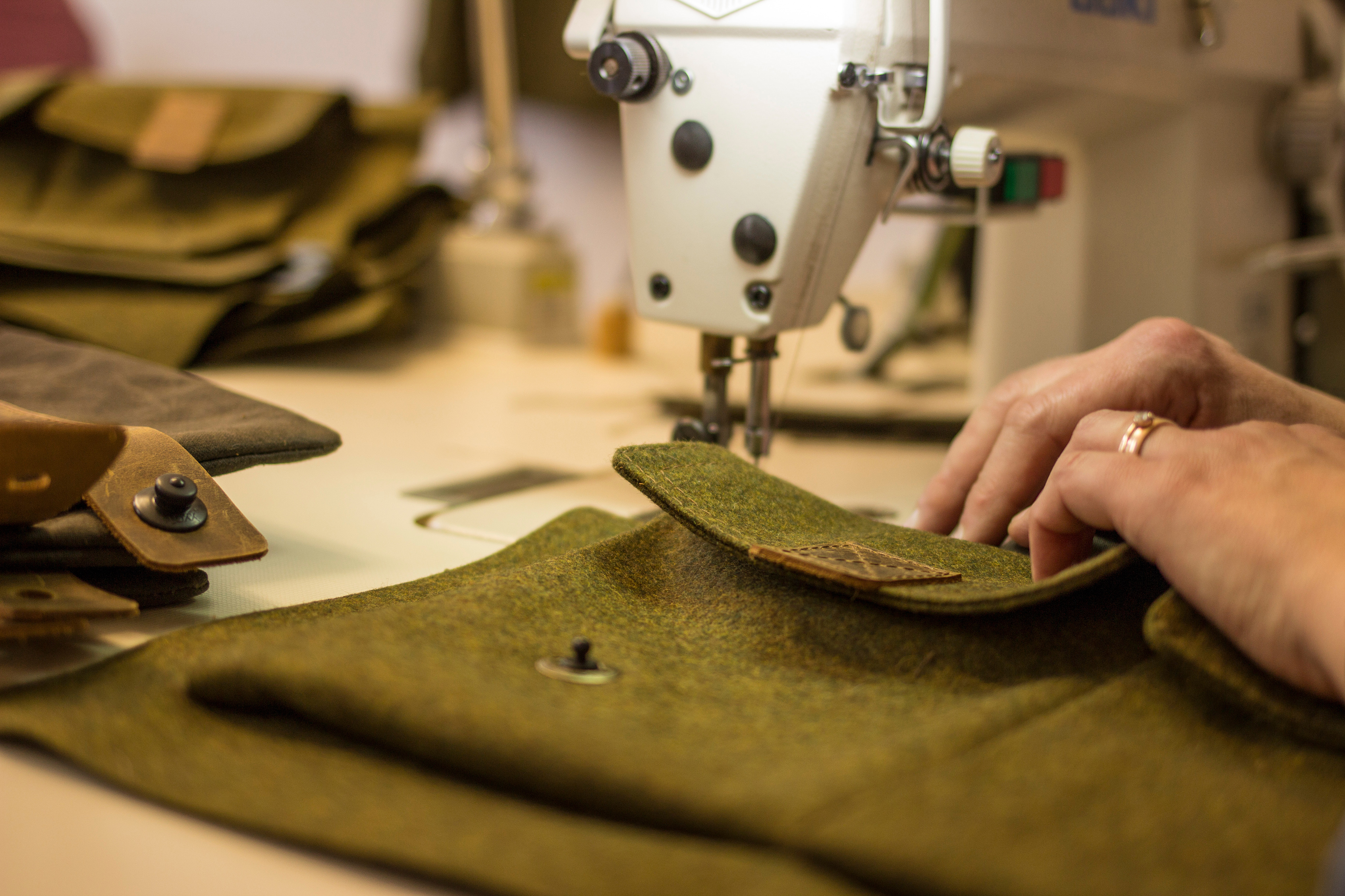 Hände einer Person nähen mit einer Nähmaschine an einer olivgrünen Stofftasche mit Lederelementen, daneben liegen fertiggestellte Taschen.
