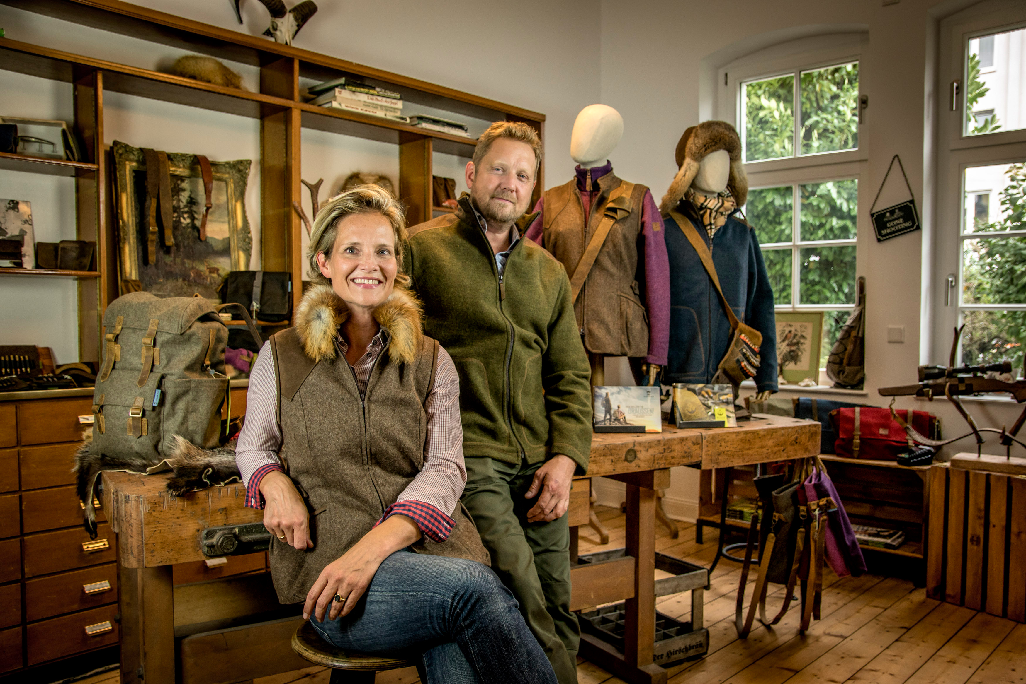 Ein Mann und eine Frau in traditioneller Kleidung stehen in einer Werkstatt mit Holzregalen, Kleidung und Accessoires, vor einem Holztisch mit Werkzeugen. Im Hintergrund sind Fenster und grüne Pflanzen durch das Glas sichtbar.