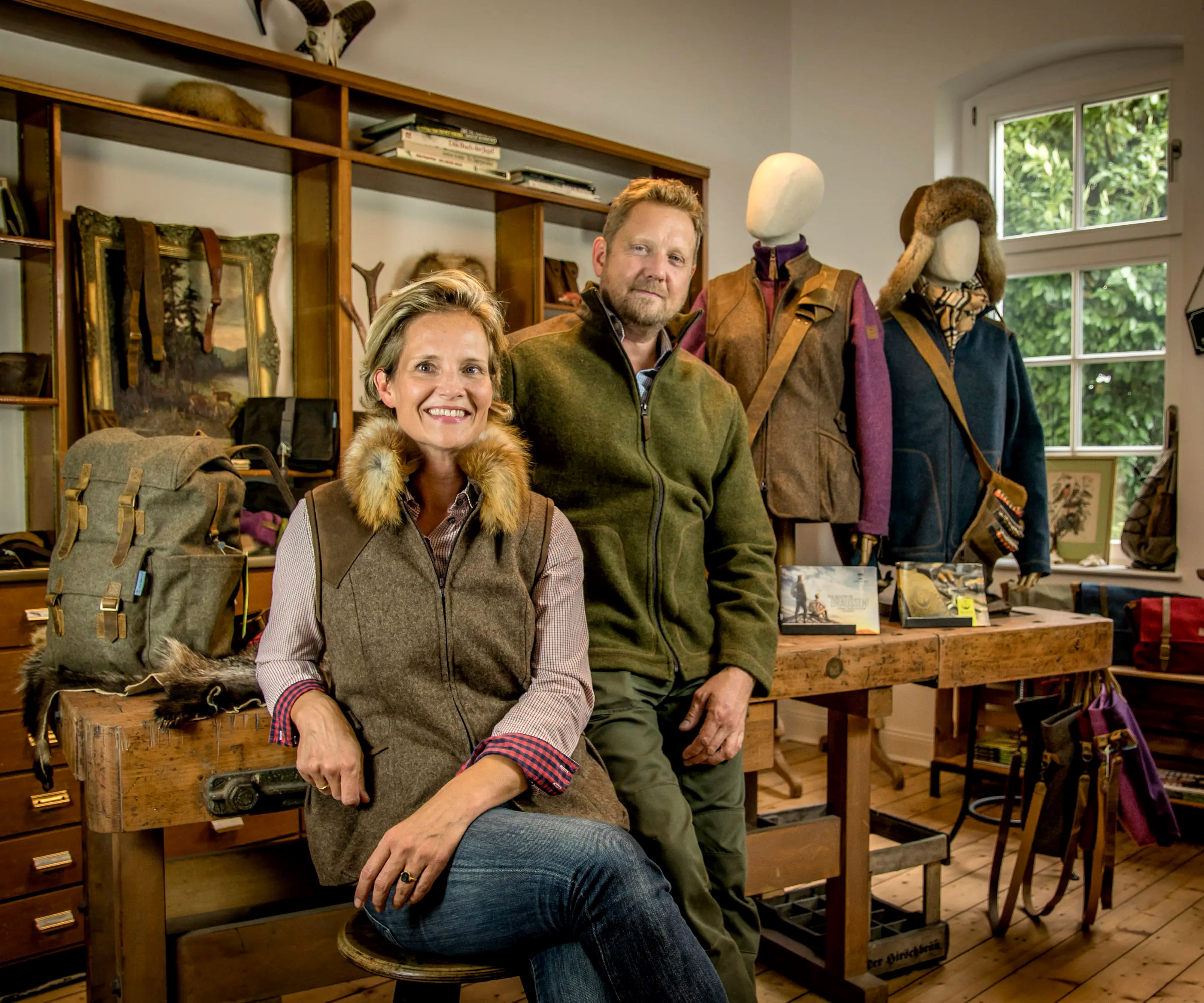 Ein Mann und eine Frau stehen in einem Laden mit Outdoor-Bekleidung. Sie lehnen an einem Holztisch, auf dem Kleidung und Accessoires ausgestellt sind. Im Hintergrund sind Regale mit Dekorationsgegenständen zu sehen. Bild verlinkt zum Herstellerportrait.