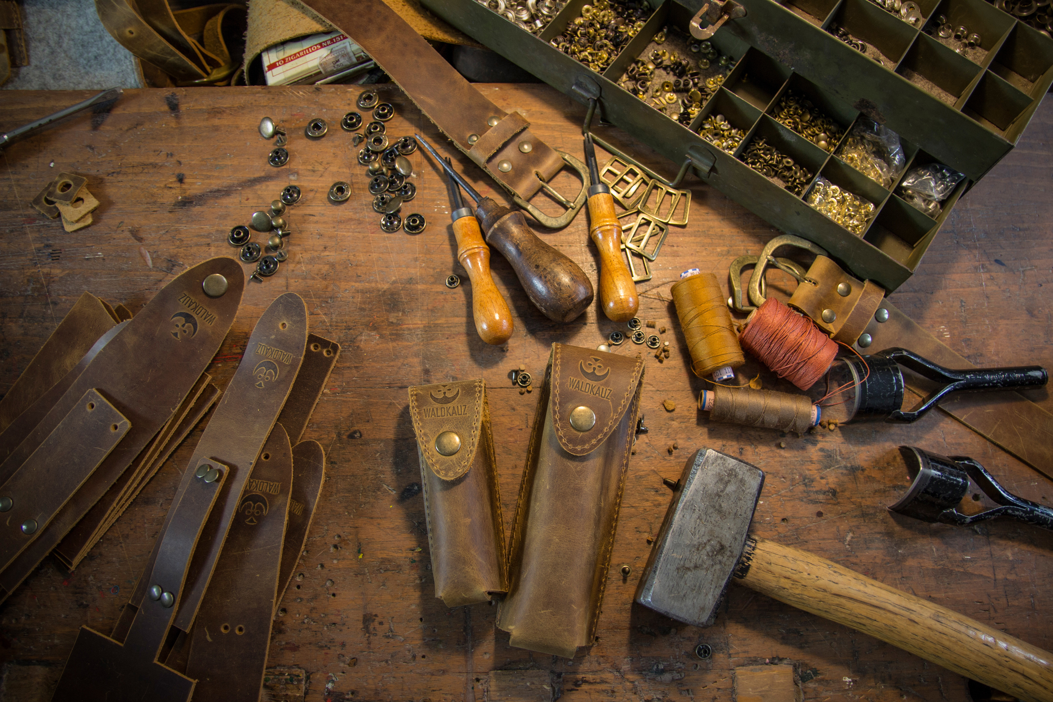 Lederwerkstatt mit braunen Ledertaschen, Werkzeugen wie Hämmern und Ahlen sowie Materialien auf einem Holztisch. Im Hintergrund offene Schublade mit Metallteilen. Alles deutet auf handwerkliche Lederbearbeitung hin.